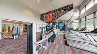 Located at 465 Memorial Dr SE in Atlanta, GA, this well-appointed fitness center offers a generously sized, single-floor exercise space designed to support a comprehensive workout experience. The room's double-height ceilings create an open, airy atmosphere, while expansive floor-to-ceiling windows along multiple walls flood the interior with natural light and provide refreshing views of the surrounding greenery. A bold, large-scale graphic mural featuring a city map with vivid red accents serves as a striking focal point on one wall, reinforcing the space's contemporary urban character. The flooring consists of patterned commercial carpet in gray and red tones that adds visual energy underfoot. The facility is comprehensively equipped with Matrix-brand cardio machines, including treadmills, ellipticals, stair climbers, and stationary bikes, as well as a full complement of strength-training equipment such as cable machines, weight benches, and resistance machines. Flat-screen televisions are mounted throughout for entertainment during workouts. A sleek spiral staircase with a dark metal railing connects to an upper mezzanine level, adding architectural interest and additional functional space. Full-length mirrors enhance the sense of depth, and the thoughtful layout ensures ample room to move freely between stations.
