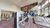 Located at 465 Memorial Dr SE in Atlanta, GA, this well-appointed fitness center offers a generously sized, single-floor exercise space designed to support a comprehensive workout experience. The room's double-height ceilings create an open, airy atmosphere, while expansive floor-to-ceiling windows along multiple walls flood the interior with natural light and provide refreshing views of the surrounding greenery. A bold, large-scale graphic mural featuring a city map with vivid red accents serves as a striking focal point on one wall, reinforcing the space's contemporary urban character. The flooring consists of patterned commercial carpet in gray and red tones that adds visual energy underfoot. The facility is comprehensively equipped with Matrix-brand cardio machines, including treadmills, ellipticals, stair climbers, and stationary bikes, as well as a full complement of strength-training equipment such as cable machines, weight benches, and resistance machines. Flat-screen televisions are mounted throughout for entertainment during workouts. A sleek spiral staircase with a dark metal railing connects to an upper mezzanine level, adding architectural interest and additional functional space. Full-length mirrors enhance the sense of depth, and the thoughtful layout ensures ample room to move freely between stations.