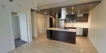 This single-floor space showcases a contemporary open-concept layout that integrates the kitchen and surrounding living areas into one cohesive environment. The kitchen serves as the centerpiece, featuring sleek dark cabinetry that contrasts sharply with light countertops on a substantial central island equipped with a built-in cooktop. A stainless steel range hood suspended from above the island adds an industrial yet refined focal point, while matching stainless steel appliances, including a full-sized refrigerator, reinforce the modern aesthetic. Upper cabinetry with under-cabinet lighting lines the walls, providing ample storage and a polished finish. Light hardwood-style flooring runs throughout, brightening the space and creating a sense of continuity between rooms. Recessed lighting is thoughtfully placed across the ceiling to ensure even illumination throughout. A hallway visible from the kitchen leads to additional rooms, hinting at a well-organized floor plan. The overall ambiance is clean, minimalist, and functional, with a neutral color palette accented by dark design elements that give the space a sophisticated, urban character.