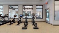 Located at 161 Grande Oaks Drive in Charleston, SC, this fitness center offers a comprehensive, thoughtfully organized workout facility designed to accommodate a wide range of training needs. The space is divided into distinct functional zones, each outfitted with professional-grade equipment. The main cardio area features a row of treadmills, ellipticals, and stationary cycling bikes, including smart-screen-equipped models, arranged along mirrored walls that amplify the sense of space and allow users to monitor their form. A dedicated central running lane with a rubberized dark track strip runs the length of the main floor, providing a clear path for movement and functional drills. The strength training section includes a multi-functional cable machine, a Smith machine, a full dumbbell rack ranging across multiple weight increments, and a red power rack suitable for barbell exercises. Additional strength machines and a flat bench round out the resistance training options. The free-motion and stretching area features hardwood flooring, yoga mats, and medicine balls, offering a quieter space for flexibility and recovery work. Throughout the facility, oversized windows and glass-panel French doors flood the interior with natural light and provide views of the surrounding greenery. Soaring ceilings fitted with large industrial-style ceiling fans and recessed lighting maintain a comfortable, airy atmosphere. Mirrors are strategically placed across multiple walls to enhance both functionality and the open feel of the space.