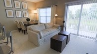 This single-floor residence showcases an open-concept layout that seamlessly connects the living and dining areas, creating a cohesive and functional shared space. The living area is anchored by a generously sized, light-colored upholstered sofa paired with dark wood cube ottomans that serve as both a coffee table and additional seating. Soft, neutral wall tones provide a warm backdrop, while framed botanical artwork adds a subtle decorative touch.

Natural light is a defining feature of this space, with multiple windows dressed in light-filtering shades and curtains that allow daylight to permeate throughout. A sliding glass door leads to an outdoor balcony, offering views of surrounding greenery and further enhancing the sense of openness. Vertical blinds frame the balcony entry, allowing for adjustable privacy.

The adjoining dining area accommodates a dark wood table with upholstered chairs, complemented by a potted topiary and a warm floor lamp that adds ambient lighting. Carpeted flooring runs throughout, contributing to a comfortable and cohesive aesthetic. The overall ambiance is calm, neutral, and well-lit, making the space feel inviting and practical.