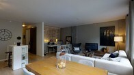 This single-floor space presents an open-concept layout that integrates multiple functional areas within a cohesive, contemporary design. The living area anchors the space with a large, plush white sectional sofa paired with a distinctive egg-style accent chair in dark upholstery, creating an inviting and stylish seating arrangement. A flat-screen television is mounted opposite the seating area, complemented by a rustic wooden coffee table that adds an organic contrast to the otherwise sleek aesthetic. A dedicated workspace or vanity area features a streamlined white desk, open shelving unit, and warm accent lighting, including a decorative backlit wall panel that lends an artistic, textured focal point. The dining area includes a light wood table with upholstered chairs, positioned to allow easy flow throughout the space. Neutral grey walls and light hardwood flooring run throughout, contributing to a calm, modern ambiance. Industrial-style concrete column details add architectural character, while recessed lighting maintains a warm, even illumination across the open floor plan.