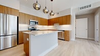 This single-floor residence showcases a thoughtfully designed open layout with a distinctly modern aesthetic. The kitchen serves as the centerpiece of the living area, featuring flat-front cabinetry in warm wood tones extending to the upper walls, providing generous storage. A sizeable central island with a crisp white countertop offers additional workspace and casual seating, while a trio of sculptural smoked-glass pendant lights hang overhead, adding a contemporary focal point. Stainless steel appliances, including a refrigerator, range, microwave, and dishwasher, complete the well-appointed space. Light-toned hardwood-style flooring runs throughout the main living areas, enhancing the sense of openness and continuity. Notably tall ceilings amplify the airy feel of the open-plan living and kitchen zones. The bedroom features plush neutral carpeting and a window fitted with horizontal blinds that allows natural light to filter in. The bathroom is finished with large-format tile flooring, a wood-toned vanity, and a full tub-shower combination. In-unit laundry facilities are also present, adding everyday convenience to this clean, modern residence.