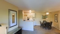This single-floor residence offers an open-concept layout that connects the main living areas in a functional and cohesive design. The kitchen is positioned at the heart of the space, featuring a peninsula-style counter with a raised breakfast bar that serves as a natural divider between the kitchen and the surrounding areas. White cabinetry lines the walls, providing ample storage, while built-in appliances including a microwave and refrigerator contribute to a clean, streamlined look. A ceiling-mounted light fixture illuminates the kitchen workspace effectively. Adjacent to the kitchen, the dining area comfortably accommodates a full dining set and is lit by a ceiling fan with integrated lighting. The living area extends naturally from the dining space, featuring plush carpeted flooring and warm, neutral-toned walls that create a welcoming ambiance throughout. A textured ceiling finish is consistent across all areas, adding a subtle design element overhead. Decorative touches, including framed wall art, enhance the residential character of the space. The overall layout is thoughtfully designed to maximize flow and connectivity between functional zones, making it a practical and comfortable living environment.