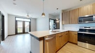 This well-appointed single-floor residence combines contemporary design with practical functionality throughout its thoughtfully arranged layout. The open-concept kitchen serves as the heart of the space, outfitted with warm wood cabinetry, a suite of stainless steel appliances including a refrigerator, electric range, built-in microwave, and dishwasher, all set against a clean white tile backsplash. A peninsula with a undermount sink and light countertops provides both additional prep space and a natural division between the kitchen and the living area. Pendant lighting overhead adds a modern touch to the space. The living area extends from the kitchen, featuring light wood-style flooring, a ceiling fan, and large glass doors that invite natural light and provide outdoor access. The bedrooms are carpeted for comfort, each featuring a ceiling fan and generous windows that frame outdoor views. The full bathroom is finished with matching wood-toned cabinetry, a white countertop vanity with dual sinks, a soaking tub with tile surround, and light wood flooring. A walk-in closet with built-in shelving connects directly to the bathroom. Consistent use of warm wood tones, neutral walls, and dark door frames creates a cohesive, modern aesthetic throughout the entire residence.