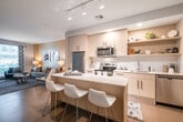 This contemporary single-floor residence offers a thoughtfully designed open-concept layout that seamlessly connects the kitchen, dining, and living areas. The kitchen is outfitted with light wood cabinetry, quartz countertops, a large center island with bar seating, and a full suite of stainless steel appliances including a refrigerator, electric range, microwave, and dishwasher. Open shelving adds a modern, airy touch alongside ample cabinet storage. Track and recessed lighting illuminates the space efficiently throughout.

The living area is warm and inviting, featuring neutral walls accented by a rich golden tone, large windows that bring in natural light and offer leafy outdoor views, and durable hard-surface flooring. The space is well-proportioned for both relaxing and entertaining.

The bedroom is a comfortable retreat, highlighted by a soft blue-grey accent wall, carpeted flooring, and a generous window overlooking lush greenery. A nearby dresser area with a round mirror adds functional charm.

The bathroom is clean and modern, finished with tile flooring, a white vanity with an integrated sink, and a tall storage column. An open doorway connects back to the main living area, reinforcing the unit's easy, flowing floor plan. Overall, the residence balances contemporary design with a warm, livable ambiance.