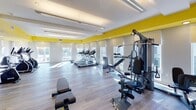 The CiTiVillage Fitness Center in Linden, NJ is a thoughtfully designed, single-floor facility that combines a vibrant aesthetic with a comprehensive range of workout equipment. The space is immediately distinguished by its bold yellow accent band running along the upper walls, set against a clean white ceiling and neutral lower walls, creating an energetic and motivating atmosphere.

The flooring throughout is a light-toned wood-style surface that lends a modern, polished look while remaining practical for fitness use. Recessed and linear LED lighting ensures the space is evenly and brightly lit, complementing the abundant natural light that pours in through multiple large windows fitted with roller shades, offering views of the surrounding area.

The cardio zone features a row of treadmills, elliptical trainers, and stationary bikes, positioned to take advantage of the natural light. A dedicated strength-training area includes a multi-station cable machine, adjustable benches, and a well-stocked dumbbell rack ranging across various weights. A full-length mirror wall spans one side of the room, visually expanding the space and aiding in proper form during workouts. The open layout provides ample room between equipment, ensuring a comfortable and functional exercise environment.