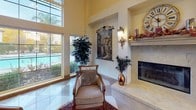 This single-story clubhouse exudes a warm, Tuscan-inspired ambiance throughout its generously proportioned interior. The main gathering area showcases soaring ceilings that amplify the sense of openness, with tall floor-to-ceiling windows flooding the space with natural light and offering views of the outdoor pool area. A striking diamond-patterned tile floor runs throughout, adding a polished, classic character to the space.

A focal point of the lounge is the stone-surround fireplace, topped with a large decorative clock and flanked by ornate wall-hung tapestries depicting Italian countryside landscapes. Arched niches in the warm golden-toned walls further enhance the Mediterranean aesthetic. Comfortable, upholstered seating areas are arranged throughout, including a cozy nook with plantation-shuttered windows ideal for intimate gatherings.

Classical columns frame the transition into a full kitchen, which features rich dark wood cabinetry, stainless steel appliances, and recessed lighting set against terra cotta-toned accent walls. The entry corridor welcomes visitors with decorative tapestry art, an ornamental potted tree, and elegant ceramic accents. Throughout the clubhouse, thoughtful décor details — from wrought-iron wall fixtures to area rugs — contribute to a refined yet inviting community gathering space.
