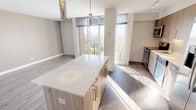 This single-floor unit showcases a contemporary open-concept layout that seamlessly connects the kitchen, dining, and living areas. The kitchen is appointed with light wood cabinetry, a tile backsplash, stainless steel appliances including a refrigerator, range, microwave, and dishwasher, and a generous island with a white quartz countertop. Stylish pendant lights hang above the island, adding a warm accent to the otherwise neutral palette. Track lighting along the kitchen ceiling provides additional task illumination.

The living area benefits from large, floor-to-ceiling windows that draw in abundant natural light and offer views of the surrounding urban landscape and greenery. The wood-style flooring, rendered in a warm medium tone, extends throughout the main living space, contributing to a cohesive and modern aesthetic. Neutral gray walls complement the light tones of the cabinetry and countertops throughout.

A bathroom is accessible via a private corridor, featuring light tile flooring, clean white walls, a wood-toned vanity with storage, and a wall-mounted mirror. The overall design of the unit emphasizes clean lines, modern finishes, and a bright, airy atmosphere enhanced by the generous window placement and carefully chosen materials.
