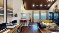 Located at 267–269 Charlotte Hwy in Asheville, NC, this single-floor space is defined by a striking architectural character that blends rustic warmth with contemporary design. The interior is anchored by a dramatic floor-to-ceiling stacked stone fireplace, which serves as a commanding focal point beneath soaring vaulted ceilings finished in rich, dark-stained wood planking. This combination of natural materials lends the space a refined yet grounded aesthetic throughout.

Dark hardwood flooring runs continuously across the open layout, connecting a comfortable lounge area to a dedicated workspace and a kitchenette. The lounge features a cozy seating arrangement with a velvet sofa, leather armchairs, and a glass coffee table, complemented by curated artwork and a statement display cabinet. Tall windows dressed with layered drapes and blinds flood the interior with natural light while maintaining privacy.

The kitchenette is well-appointed with granite countertops, dark wood cabinetry, bar-height seating, and a coffee station, backed by a large-scale mountain landscape mural that adds a bold artistic touch. Recessed and pendant lighting throughout ensures a warm, well-lit ambiance. The overall atmosphere is inviting and thoughtfully curated, reflecting the creative spirit of the Asheville region.