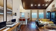 Located at 267–269 Charlotte Hwy in Asheville, NC, this single-floor space is defined by a striking architectural character that blends rustic warmth with contemporary design. The interior is anchored by a dramatic floor-to-ceiling stacked stone fireplace, which serves as a commanding focal point beneath soaring vaulted ceilings finished in rich, dark-stained wood planking. This combination of natural materials lends the space a refined yet grounded aesthetic throughout.

Dark hardwood flooring runs continuously across the open layout, connecting a comfortable lounge area to a dedicated workspace and a kitchenette. The lounge features a cozy seating arrangement with a velvet sofa, leather armchairs, and a glass coffee table, complemented by curated artwork and a statement display cabinet. Tall windows dressed with layered drapes and blinds flood the interior with natural light while maintaining privacy.

The kitchenette is well-appointed with granite countertops, dark wood cabinetry, bar-height seating, and a coffee station, backed by a large-scale mountain landscape mural that adds a bold artistic touch. Recessed and pendant lighting throughout ensures a warm, well-lit ambiance. The overall atmosphere is inviting and thoughtfully curated, reflecting the creative spirit of the Asheville region.