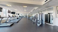 Innova Fitness Center in Cleveland, Ohio offers a comprehensive, professionally designed fitness facility spread across a single, expansive floor. The space is defined by its clean, modern aesthetic, featuring a drop-panel ceiling with recessed and flush lighting that keeps the entire floor plan bright and energizing throughout the day. Large floor-to-ceiling windows line the perimeter, flooding the interior with natural light and providing open views of the surrounding Cleveland neighborhood, creating an inviting atmosphere for workouts. The main workout floor is finished with durable rubber flooring, ideal for high-traffic fitness use, and is divided into distinct functional zones. The cardio section is well-stocked with a full row of treadmills, ellipticals, and stationary bikes, many positioned directly in front of wall-mounted flat-screen televisions for an engaging exercise experience. The strength training area features a wide selection of cable machines, selectorized weight machines, and functional training rigs, including a multi-cable cross system. Additional accessories such as stability balls and resistance equipment are neatly organized along mirrored walls, which amplify the sense of space. A dedicated open-floor zone provides ample room for stretching, functional movement, and bodyweight training. The overall ambiance is clean, spacious, and professional, making Innova Fitness Center a well-rounded facility suited for diverse fitness goals.
