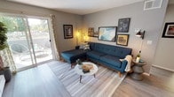 This thoughtfully designed single-floor residence showcases a cohesive mid-century modern aesthetic throughout its well-organized layout. The living room serves as the heart of the space, featuring light wood-style flooring, a tufted teal sectional sofa with walnut legs, and a round marble-top coffee table. A large sliding glass door floods the room with natural light and opens onto a private balcony, offering outdoor views and additional lounging space.

Adjacent to the living area, the dining space is distinguished by a round wood-grain table paired with upholstered chairs, a ceiling fan overhead, and a built-in wet bar complete with a sink, granite-style countertop, white cabinetry, and wine glass storage — an ideal feature for entertaining. A trio of gold-framed mirrors adds visual depth to the area.

The kitchen is efficiently designed with white shaker cabinets, granite-style countertops, and stainless steel appliances including a range and refrigerator, all complemented by the same light wood flooring found throughout.

The bedroom offers plush carpeting, a sliding-door closet, and a natural light source through a window with blinds and drapes. The full bathroom features a single-sink vanity with granite-style countertop, a soaking tub with shower, and wood-look flooring.