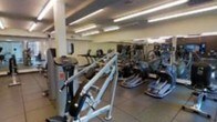 This fitness center offers a comprehensive workout environment designed to accommodate a variety of exercise routines. The space is laid out across a single floor, with an open floor plan that allows for easy navigation between equipment stations. A robust selection of cardio machines takes center stage, including treadmills, ellipticals, stair climbers, and stationary bikes, all arranged to maximize accessibility and flow throughout the room. Strength training equipment and additional machines line the perimeter, providing users with a full range of workout options. The facility is brightly illuminated by rows of fluorescent overhead lighting, ensuring excellent visibility across the entire space. The flooring features durable tile, well-suited for a high-traffic fitness environment. Structural columns are integrated into the layout without impeding movement or access to equipment. The overall ambiance is functional and professional, reflecting a space purpose-built for fitness with an emphasis on variety and convenience. Mirrored wall sections enhance the sense of openness and allow users to monitor their form during exercise. The space is clean, organized, and equipped to support both casual and dedicated fitness routines.