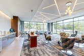 Located in Lynn, MA, this impressive lobby spans a generous single floor, offering a thoughtfully designed and welcoming environment. The entrance features large-format porcelain tile flooring and a striking wood slat ceiling detail that adds warmth and texture to the space. A cozy booth seating nook tucked beneath the slatted wood canopy provides an intimate spot for small gatherings, complemented by compact café-style tables and blue accent chairs.

Moving through the lobby, the space opens into a bright and airy lounge area bathed in natural light from expansive floor-to-ceiling glass windows that frame views of the surrounding greenery. Geometric linear LED lighting is embedded into the white ceiling in an angular pattern, creating a modern and dynamic visual overhead. An assortment of stylishly upholstered swivel chairs, lounge seating, and a rich wood console table furnish this area, fostering a comfortable and professional atmosphere.

A separate seating alcove features a coffered circular ceiling detail adorned with a sculptural pendant light, with curated coastal-themed artwork displayed on the walls. A glass-enclosed interior office space, accessible via dark-framed doors, adds functional versatility. The lobby also includes a vending area and convenient amenities, making it a well-rounded, inviting hub for residents and visitors alike.