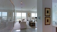 This single-floor space showcases a thoughtfully designed living area characterized by a clean, contemporary aesthetic. The room is anchored by large floor-to-ceiling windows that span multiple walls, flooding the interior with abundant natural light and framing sweeping panoramic views of the surrounding landscape and skyline. The elevated positioning of the space enhances these vistas, making them a defining architectural feature.

The living area is furnished in a cohesive neutral palette, featuring a plush sofa adorned with coordinating cushions, a skirted accent chair, and additional seating that encourages an open, conversational layout. A low-profile wooden coffee table with open shelving sits atop a light-toned area rug, grounding the seating arrangement. Subtle decorative elements, including abstract wall art and table lamps, complement the refined atmosphere without overwhelming the minimalist design.

The high, smooth ceilings are equipped with recessed track lighting, providing flexible illumination throughout the space. The polished tile flooring reflects the natural light, amplifying the sense of openness and airiness. A smart home control panel is visible on one wall, indicating modern technological integration. Overall, the space exudes a calm, sophisticated ambiance suited for comfortable, elevated living.