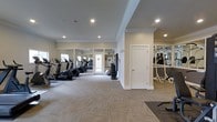 This fitness center offers a spacious and well-organized workout environment designed to accommodate a wide range of exercise needs. The layout is thoughtfully divided into distinct zones, including a dedicated cardio area lined with treadmills and elliptical machines, a free weight station with a full dumbbell rack, and a strength training section featuring cable and resistance equipment. Large mirrors span the walls throughout, creating a sense of openness while allowing users to monitor their form during workouts. The flooring consists of durable, textured carpet tile that provides both cushioning and traction underfoot. Recessed lighting illuminates the space evenly from above, complemented by elegant crown molding that adds a refined architectural touch to the room's clean, neutral palette of beige and white. A glazed door at the far end of the cardio area allows natural light to filter in and offers a view to an outdoor area beyond. The overall ambiance is bright, clean, and functional, striking a balance between practicality and a polished aesthetic that makes for a welcoming environment for daily fitness routines.