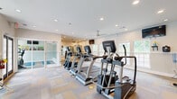 Located at 6687 Skipper Rd in Macon, Georgia, this fitness center offers a well-organized and fully equipped workout facility across a single floor. The space is designed with a clean, bright aesthetic, featuring neutral-toned walls, patterned carpet tile flooring in soft beige and tan hues, and recessed lighting throughout that ensures consistent illumination across the entire area.

The main workout area is outfitted with a comprehensive selection of True-brand cardio equipment, including treadmills, elliptical trainers, and stationary bikes, arranged in a row along one side of the room. Opposing this section is a dedicated strength training zone featuring multi-function cable machines, a weight bench, and additional resistance equipment. Large wall-mounted mirrors in the strength area enhance the sense of space and allow users to monitor their form during workouts.

Natural light pours in through multiple large, white-framed windows that line the walls, offering pleasant outdoor views of the surrounding greenery. Ceiling fans are installed throughout to maintain comfortable airflow. Wall-mounted flat-screen televisions are positioned for easy viewing from the cardio equipment. A water cooler station is conveniently available for hydration. Glass-panel doors provide access to an adjacent area, adding to the open and connected feel of the facility.