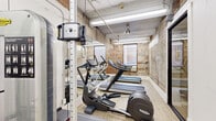 Located at 410 W 7th Street in the heart of Fort Worth, Texas, this fitness center offers a well-equipped workout space with a distinct industrial character. The room is defined by its exposed brick walls, which lend a raw, authentic aesthetic that contrasts beautifully with the light wood-look flooring underfoot. The ceiling features visible ductwork and overhead fluorescent lighting, reinforcing the industrial design sensibility throughout.

The space is outfitted with a range of Technogym cardio machines, including multiple treadmills and elliptical trainers, arranged efficiently along the length of the room. A multi-functional cable and weight machine provides strength training options, making this a versatile facility for varied workout routines. Full-length mirrors line one wall, visually expanding the space and aiding in form correction during exercise.

Natural light filters in through a window at the far end of the room, offering a view of the surrounding brick urban streetscape, adding an open and airy quality to the otherwise compact environment. Entry is through sleek, black-framed glass double doors that maintain visual transparency and connectivity to the adjoining corridor. The overall atmosphere is functional yet stylish, blending the building's historic architectural bones with modern fitness amenities.