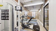 Located at 410 W 7th Street in the heart of Fort Worth, Texas, this fitness center offers a well-equipped workout space with a distinct industrial character. The room is defined by its exposed brick walls, which lend a raw, authentic aesthetic that contrasts beautifully with the light wood-look flooring underfoot. The ceiling features visible ductwork and overhead fluorescent lighting, reinforcing the industrial design sensibility throughout.

The space is outfitted with a range of Technogym cardio machines, including multiple treadmills and elliptical trainers, arranged efficiently along the length of the room. A multi-functional cable and weight machine provides strength training options, making this a versatile facility for varied workout routines. Full-length mirrors line one wall, visually expanding the space and aiding in form correction during exercise.

Natural light filters in through a window at the far end of the room, offering a view of the surrounding brick urban streetscape, adding an open and airy quality to the otherwise compact environment. Entry is through sleek, black-framed glass double doors that maintain visual transparency and connectivity to the adjoining corridor. The overall atmosphere is functional yet stylish, blending the building's historic architectural bones with modern fitness amenities.