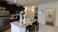 This well-appointed single-floor residence offers a thoughtfully designed open-concept layout that seamlessly connects the kitchen and living areas. The kitchen is a standout feature, showcasing rich dark espresso cabinetry that extends to the upper walls, providing ample storage while creating a bold, contemporary contrast against the light-toned walls and flooring. A generous granite-topped kitchen island serves as both a prep space and a casual dining area, accompanied by sleek black leather bar stools. Elegant pendant lights hang above the island, complementing the track lighting overhead to create a warm, well-lit environment. Stainless steel appliances, including a built-in microwave and a range, add a polished, modern touch to the kitchen. Neutral-toned carpeting flows through the adjoining living spaces, contributing to a clean and cohesive aesthetic. A glimpse into the adjacent areas reveals a neatly organized workspace or den with open shelving, and a framed wall feature that adds a decorative accent. The overall ambiance is modern and inviting, with a balanced mix of dark and light tones, purposeful lighting, and functional design elements that make this space both practical and visually appealing.