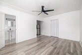 This 230 square foot studio apartment in Los Angeles offers a thoughtful blend of compact living and modern design. The space is efficiently laid out on a single floor, comprising a living room, kitchen, and bathroom. The living area features light-toned wood-look flooring that extends throughout, creating a sense of continuity and spaciousness. A modern ceiling fan with sleek blades adds both style and functionality.

The kitchen, though compact, is well-equipped with white cabinetry, providing ample storage. It includes a stainless steel refrigerator and a dark countertop, creating a striking contrast against the white cabinets. A sink with a modern black faucet completes the functional space.

The bathroom, while not expansive, offers essential amenities including a shower with a black showerhead, maintaining the contemporary aesthetic found throughout the apartment. The overall color scheme of whites and light neutrals contributes to a bright, airy feel despite the limited square footage.

Crown molding adds a touch of elegance to the space, while the minimalist design approach maximizes the available area. This studio apartment represents an ideal solution for those seeking efficient, low-maintenance urban living in the heart of Los Angeles.