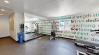 This single-floor fitness center is designed to provide residents with a functional and motivating workout environment. The space features durable dark rubber flooring throughout, providing a practical and slip-resistant surface suited for exercise. A standout design element is the bold motivational typography wall mural, which displays fitness-themed words and phrases in white and teal across multiple walls, creating an energetic and encouraging atmosphere.

The gym is equipped with a selection of cardio machines, including treadmills and an elliptical trainer, positioned to allow comfortable use. A multi-function cable weight machine offers strength training options. A wall-mounted flat-screen television is positioned for easy viewing during workouts. Floor-to-ceiling mirrors line one wall, visually expanding the space while allowing users to monitor their form.

Natural light filters in through windows and a glass-panel door, which provides direct access to an outdoor area. Recessed lighting in the textured ceiling supplements the natural light, keeping the space bright and well-lit. A separate entry area adjacent to the fitness floor includes a bank of resident mailboxes and a recycling bin, indicating this fitness center serves as a shared amenity within a larger residential community. The overall layout is open and efficiently organized for a streamlined workout experience.