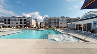 This outdoor amenity area in Sumter, South Carolina, is designed to offer a resort-like experience within a well-appointed residential community. The centerpiece is a generously sized swimming pool with crystal-clear blue water, complemented by an adjacent splash zone featuring built-in water features — ideal for leisure and recreation. The pool deck is finished in light-toned concrete, providing ample space for sunbathing and socializing, and is furnished with modern sling-style lounge chairs and shade umbrellas in deep navy blue.

Surrounding the pool area, the landscaping is thoughtfully curated, incorporating palm trees, flowering rose bushes, and mulched garden beds that add color and a subtropical character fitting for the South Carolina climate. A black aluminum fence lines portions of the perimeter, framing manicured lawn areas and maintaining an open yet defined boundary.

A clubhouse-style structure with a cupola roofline anchors one side of the pool area, providing architectural interest. The broader community features multi-story residential buildings clad in a combination of blue and neutral siding with stone accents and dormer windows. Together, the outdoor spaces create a bright, welcoming ambiance well-suited to the warm Southern climate.