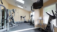Located at 397 Brixton Drive in Virginia Beach, Virginia, this single-floor amenity space is thoughtfully designed to support an active lifestyle. The entryway greets visitors with warm-toned slate-style tile flooring and neutral walls accented by framed nautical artwork, setting a welcoming coastal ambiance. A glass entry door allows natural light to pour into the corridor, which provides easy access to the facility's key areas and includes safety features such as a clearly mounted fire alarm system.

The centerpiece of the space is a well-appointed fitness room outfitted with rubber flooring for comfort and durability. The room features a multi-function cable machine, a treadmill, and an elliptical trainer, catering to a range of workout preferences. A large wall-mounted flat-screen television provides entertainment during exercise sessions, while a full-length mirror wall visually expands the space and aids in form monitoring. Skylights overhead flood the room with natural light, complementing the window treatments that allow for adjustable brightness. A small utility shelf with sanitizing supplies ensures the space remains clean and functional.

Overall, the space strikes a balance between practicality and comfort, offering a focused fitness environment with thoughtful details throughout.