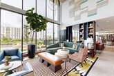 Located at 14020 Stream Way in Charlotte, NC, this sophisticated single-floor living space offers a generously proportioned communal lounge designed with a bold, contemporary aesthetic. The space is anchored by dramatic double-height ceilings that amplify the sense of openness, while expansive floor-to-ceiling windows flood the interior with natural light and frame views of the outdoor terrace and surrounding community.

The living area is thoughtfully divided into distinct seating zones, each curated with eclectic, design-forward furnishings. A curved sage-green sectional sofa, patterned armchairs, and layered geometric rugs create a warm yet vibrant atmosphere. Striking teal and neutral wall art panels punctuate the soaring white walls above, adding an artistic focal point.

A sleek built-in fireplace clad in metallic tile serves as a dramatic feature wall, paired with a mounted flat-screen display and flanked by floor-to-ceiling wood-paneled doors that open directly to the outdoor pool area. Opposite, a fully appointed wet bar with brass-accented cabinetry, mirrored shelving, and counter seating provides an elegant entertaining hub.

Rich wood-paneled walls, built-in bookshelves with curated décor, herringbone hardwood flooring, and a bold checkered tile inset contribute to the space's layered, upscale character. The overall ambiance is lively yet refined, blending comfort with high-design sensibility.