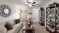 This single-story residence offers a thoughtfully arranged floor plan that balances communal living spaces with private retreats. The living room serves as the heart of the home, featuring plush carpet, a ceiling fan, and an open sightline that connects seamlessly to the dining area and kitchen. A decorative shelving unit and tasteful wall accents contribute to the room's welcoming character, while a front-facing window allows natural light to filter through.

The kitchen is designed for practicality, featuring warm oak cabinetry extending to the upper walls for generous storage, tile flooring, a gas range, and a full-size refrigerator. A breakfast bar connects the kitchen to the dining area, which accommodates a full dining set and transitions naturally into the living space.

The bedrooms are carpeted and equipped with ceiling fans. The primary bedroom offers a quiet, neutral palette, while additional bedrooms provide comfortable accommodations with access to walk-in or reach-in closets. Each of the two bathrooms features an oak vanity with ample drawer storage, a tub-shower combination, tile flooring, and a well-lit mirror above the sink, providing functional and clean finishing details throughout.