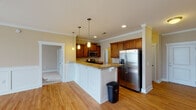 Located at 7205 Wrightsville Ave in Wilmington, NC, this single-floor residence offers a well-appointed layout designed for comfortable living. The kitchen serves as a central highlight, featuring rich wood cabinetry, granite countertops with warm gold tones, stainless steel appliances including a refrigerator with ice dispenser and built-in microwave, and a spacious white island with pendant lighting overhead. Hardwood flooring flows throughout the main living and kitchen areas, adding warmth and continuity to the space. The living area is distinguished by elegant wainscoting along the lower walls, crown molding, a ceiling fan, and a large multi-pane window that floods the room with natural light. A dedicated laundry area with washer and dryer hookups is conveniently tucked adjacent to the kitchen. The bedrooms are carpeted for comfort, with one featuring a ceiling fan and access to a walk-in closet outfitted with wire shelving. The bathrooms include a full tub-shower combination as well as a separate glass-enclosed standing shower, offering practical flexibility. Neutral warm tones throughout the home create a cohesive and inviting ambiance suited to a variety of décor styles.