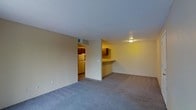 This single-floor unit offers an open-concept layout connecting the living area to the kitchen. The living space features wall-to-wall carpeting in a neutral gray tone, providing a comfortable and cohesive foundation throughout. Smooth, light-colored walls contribute to a clean, understated aesthetic, while a textured ceiling adds subtle visual character.

A partial wall with a pass-through counter separates the living area from the kitchen, creating a functional division while maintaining an open feel. The kitchen is equipped with dark wood cabinetry offering ample storage, and includes a full-size refrigerator visible through the adjoining doorway. A ceiling-mounted light fixture in the kitchen casts a warm ambient glow, complementing the overall warmth of the space.

The layout is straightforward and practical, with a clearly defined transition between the main living zone and the kitchen area. An HVAC vent is visible near the ceiling, indicating climate control throughout. The overall ambiance is simple and functional, suited to everyday living with an efficient use of space.