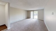 This two-bedroom, one-bathroom apartment in Beaverton, Oregon offers a practical and comfortable single-floor layout. The living room is generously proportioned, featuring plush carpeting, a baseboard heating system, and a sliding glass door with vertical blinds that opens to an outdoor view, filling the space with natural light. The combined dining and kitchen area is well-appointed with white cabinetry, dark countertops, white appliances including a range, dishwasher, and refrigerator, as well as hardwood-style flooring and a window above the sink. A dedicated laundry area houses a stacked washer and dryer unit, adding everyday convenience. The primary bedroom is a comfortable size, with plush carpeting, a baseboard heater, a window for natural light, and a double-door closet providing ample storage. The second bedroom also features carpeting, a baseboard heater, and a window. The bathroom includes a full tub and shower combination with clean white finishes. Textured ceilings are consistent throughout the unit, and neutral warm-toned walls create a cohesive, move-in ready atmosphere. An entrance and hallway connect the rooms efficiently, completing this well-organized and functional home.