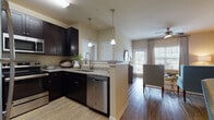 This single-floor residence is designed with a cohesive, contemporary aesthetic carried throughout each room via warm neutral walls, wood-look flooring, and rich dark-espresso cabinetry. The kitchen serves as a central hub, featuring granite countertops, stainless steel appliances including a refrigerator, microwave, and dishwasher, and a peninsula with pendant lighting overhead that opens directly to the living and dining areas. This open-concept layout promotes a connected, airy feel. The dining area comfortably accommodates a round table and sits adjacent to the living room, which receives natural light through large windows dressed with blinds and draping curtains. Ceiling fans are present in the living space and each bedroom, contributing to comfort throughout. The bedrooms are well-proportioned, each featuring large windows that allow ample natural light and include closet storage. The two bathrooms are consistently finished with granite-topped dark vanities, undermount sinks, wood-look flooring, and tub-shower combinations. One bathroom connects to a walk-in closet, adding practical storage. The overall ambiance is polished and modern with a warm, residential character.