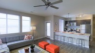 This single-floor space offers a well-designed open-concept layout that seamlessly integrates the living area and kitchen into one cohesive environment. The living room features warm hardwood-style flooring, neutral gray walls, and a ceiling fan overhead, creating a comfortable and functional atmosphere. Large windows fitted with white blinds allow abundant natural light to fill the space, enhancing its airy feel. Vibrant orange ottomans paired with a gray sofa and accent pillows add bold pops of color, giving the interior a modern and lively character. A glass-top coffee table and a tall floor lamp complement the seating arrangement. The kitchen is defined by a raised breakfast bar with metal bar stools, providing a casual dining option. Dark cabinetry contrasts elegantly with the light countertops, while stainless steel appliances — including a microwave, range, and hood — lend the kitchen a sleek, contemporary look. A subway tile backsplash adds a refined detail to the cooking area. A pendant light above the bar further enhances the kitchen's visual appeal. A door at the far end of the living space leads to an exterior entry, adding convenience and additional light to the overall layout. The space strikes a balance between modern design and everyday practicality.