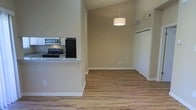 This single-floor residence offers a well-designed open-concept layout that seamlessly connects the kitchen and living areas. The kitchen is equipped with black appliances, including a refrigerator and a gas range, complemented by a stainless steel over-the-range microwave. A raised pass-through counter separates the kitchen from the adjoining living space, adding both function and visual definition to the layout. The living area benefits from soaring vaulted ceilings that amplify the sense of space and openness, while a stylish drum pendant light fixture serves as a central focal point overhead. Warm-toned, hardwood-style flooring extends throughout the main living area, adding texture and a cohesive aesthetic to the space. Neutral beige walls provide a versatile backdrop suited to a variety of décor styles. HVAC vents are integrated into the upper walls, ensuring efficient climate control throughout. A hallway visible from the main living area leads to additional spaces, including what appears to be a closet. Clean white baseboards and trim offer a crisp contrast to the warm tones of the floors and walls, completing the polished, move-in-ready feel of this inviting residence.