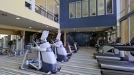 This fitness center offers a comprehensive workout environment designed to accommodate a wide range of exercise needs. The space is anchored by a generous row of cardio machines, including ellipticals and steppers, arranged across a striped, cushioned flooring that adds both style and comfort underfoot. Strength training equipment and additional cardio machines are visible toward the rear of the facility, providing ample options for a full-body workout routine.

The interior design is defined by bold navy blue walls that lend a clean, modern aesthetic throughout. Large floor-to-ceiling windows along the upper level allow natural light to pour into the space, creating an open and energizing atmosphere. Recessed ceiling lighting ensures the facility is well-lit throughout all hours of the day. The multi-story building architecture is visible through the expansive glass panels, adding a sense of depth and scale to the environment.

The layout is thoughtfully organized, allowing for easy navigation between equipment zones. Mirrors are strategically placed to enhance the sense of space and support users during their workouts. Overall, this fitness center combines a contemporary design with a well-curated selection of equipment, making it a functional and visually appealing space for health and wellness.