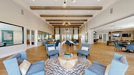 Located at 1851 Encantado Way in Kissimmee, Florida, this single-floor community clubhouse offers a generously sized, open-concept gathering space designed with both comfort and sophistication in mind. The interior is anchored by soaring ceilings adorned with exposed decorative wood beams, which lend a warm, contemporary character throughout. Light wood-look flooring runs seamlessly across the entire space, enhancing the bright, airy atmosphere created by abundant natural light pouring in through large windows and glass-paneled entry doors.

The main lounge area serves as the social centerpiece, featuring multiple seating arrangements with plush blue accent chairs, sofas, and a geometric wood coffee table accented by an elegant sculptural heron centerpiece. Statement pendant chandeliers with globe fixtures add a refined finishing touch overhead. A wall-mounted flat-screen television and textured accent wall define a secondary lounge zone, complemented by curated artwork and shiplap wall detailing.

A built-in refreshment station with cabinetry, open shelving, and a coffee setup is tucked beside a large interior glass window, offering views into an adjacent private office or meeting area. A game and dining zone features a pool table, round dining tables, and a bar-height seating area, making this clubhouse a well-rounded amenity space suited for both relaxation and recreation.