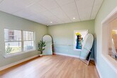 Located in Valdosta, Georgia, this single-room wellness studio is thoughtfully designed to provide a calm and functional environment for yoga, stretching, and light fitness activities. The space features smooth light hardwood flooring that extends across the entire open floor plan, creating a clean and inviting surface ideal for movement and exercise.

The walls are painted in a soft sage green, lending the room a tranquil, spa-like ambiance that promotes relaxation and focus. A wall-mounted ballet barre runs along one side, offering support for stretching and barre-style workouts. Yoga mat storage — a wall-mounted rack neatly holding rolled mats — is conveniently positioned near the entry doors, keeping the room organized and ready for use.

A large double-hung window floods the space with natural light and offers views of a landscaped outdoor area, enhancing the room's open and airy feel. Interior pass-through windows connect visually to an adjacent fitness area equipped with cardio equipment, providing a sense of spatial continuity. A full-length arched mirror adds depth to the room while supporting movement-based activities.

The drop ceiling with recessed lighting provides even, consistent illumination throughout. Two white panel doors provide access points, contributing to the room's clean, streamlined aesthetic. Overall, this studio offers a focused, peaceful setting well-suited for wellness and fitness activities.