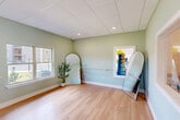 Located in Valdosta, Georgia, this single-room wellness studio is thoughtfully designed to provide a calm and functional environment for yoga, stretching, and light fitness activities. The space features smooth light hardwood flooring that extends across the entire open floor plan, creating a clean and inviting surface ideal for movement and exercise.

The walls are painted in a soft sage green, lending the room a tranquil, spa-like ambiance that promotes relaxation and focus. A wall-mounted ballet barre runs along one side, offering support for stretching and barre-style workouts. Yoga mat storage — a wall-mounted rack neatly holding rolled mats — is conveniently positioned near the entry doors, keeping the room organized and ready for use.

A large double-hung window floods the space with natural light and offers views of a landscaped outdoor area, enhancing the room's open and airy feel. Interior pass-through windows connect visually to an adjacent fitness area equipped with cardio equipment, providing a sense of spatial continuity. A full-length arched mirror adds depth to the room while supporting movement-based activities.

The drop ceiling with recessed lighting provides even, consistent illumination throughout. Two white panel doors provide access points, contributing to the room's clean, streamlined aesthetic. Overall, this studio offers a focused, peaceful setting well-suited for wellness and fitness activities.