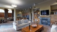 This single-floor space combines comfort and classic design in a welcoming, open-concept layout. The living area serves as the heart of the space, anchored by a handsome tile-surround fireplace that adds warmth and a refined focal point. A rich hardwood coffee table and plush upholstered sofa are arranged atop a patterned area rug, creating an inviting and well-composed seating arrangement. Adjacent to the fireplace, a built-in wood shelving unit provides tasteful display space for décor and media, complemented by a wall-mounted flat-screen television. Recessed and pendant lighting throughout ensures the space is well-lit with a warm ambiance. Structural columns add an architectural element that subtly defines zones within the open floor plan. The kitchen is efficiently designed and features stainless steel appliances, including a full-size refrigerator, and warm wood cabinetry. A raised breakfast bar with seating bridges the kitchen and living areas, making it ideal for casual dining or entertaining. Crown molding along the ceilings adds a polished, finished detail that elevates the overall aesthetic. The space presents a cohesive blend of traditional warmth and functional design throughout.