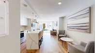 Located at 1016 Nelson Street in the heart of Vancouver's West End, this Social Room is a thoughtfully designed communal amenity space that seamlessly blends a modern kitchen with a relaxed living area. The interior features warm light hardwood flooring throughout, recessed lighting, and a textured ceiling that adds subtle depth to the room.

The kitchen is well-appointed with stainless steel appliances — including a full-size refrigerator, electric range, microwave, and dishwasher — complemented by crisp white upper cabinetry, natural wood lower accents, and a stone tile backsplash. A striking marble-veined quartz island with bar seating serves as the centerpiece, illuminated by a trio of contemporary pendant lights, making it ideal for casual gatherings.

The open living area is furnished with comfortable seating, including sofas and armchairs arranged around a marble-topped coffee table, all accented by curated artwork on the walls. Large sliding glass doors flood the space with natural light and open directly onto a spacious outdoor terrace. The terrace is equipped with a professional-grade stainless steel BBQ station and ample patio seating, set against a striking urban backdrop — perfect for outdoor entertaining. A mini-split climate system ensures year-round comfort indoors.