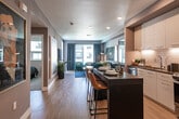 This thoughtfully designed single-floor residence blends contemporary style with functional living across a cohesive open-concept layout. Light wood flooring runs throughout the main living areas, complemented by cool-toned gray walls that lend a calm, modern ambiance.

The kitchen is a standout feature, outfitted with floor-to-ceiling white cabinetry, dark wood countertops, stainless steel appliances including a gas range, built-in microwave, dishwasher, and refrigerator, and a geometric tile backsplash. A dark-topped island with bar seating serves as both a dining and gathering space, bridging the kitchen and the open living area.

The living room is bathed in natural light from large windows and a glass balcony door, offering outdoor views. Dark velvet accent chairs, a large-format TV, and a rustic wood media console create an inviting and curated atmosphere.

The bedroom features a bold geometric accent wall, plush carpeting, and generous window views, providing a comfortable and stylish retreat. A large gold-framed mirror in the hallway adds an artistic touch to the entryway corridor.

The bathroom is clean and contemporary, featuring a glass-enclosed walk-in shower with large-format tile, warm wood-toned wall panels, and a sleek vanity with an under-mount sink.