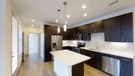 Located at 6963 E 109th Ave in Westminster, Colorado, this single-floor space showcases a well-designed, contemporary interior. The kitchen serves as a central highlight, featuring rich dark espresso cabinetry that extends floor to ceiling, offering generous storage throughout. Crisp white countertops provide a striking contrast against the dark cabinetry, while a subway tile backsplash adds a clean, classic touch to the overall aesthetic.

A central kitchen island with a white countertop surface provides additional workspace and serves as a natural gathering point. Stainless steel appliances, including a French door refrigerator with ice and water dispenser, a range with oven, over-the-range microwave, and dishwasher, contribute to the kitchen's modern, functional character.

Pendant lighting hangs above the island, complementing the recessed ceiling lights that illuminate the space evenly. Light hardwood-style flooring runs throughout, adding warmth and continuity. A hallway visible from the kitchen leads to additional areas of the space, suggesting a practical and well-organized layout. The overall ambiance is clean, modern, and move-in ready.