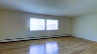 This single-floor space in the Eastgate area offers a clean, straightforward layout designed for practical use. The main living area is characterized by gleaming hardwood floors that reflect natural light, lending warmth and brightness to the interior. Two large, grid-paned windows are positioned along one wall, allowing generous amounts of daylight to fill the room while providing views of the surrounding exterior environment. The windows also contribute to a sense of openness, making the space feel airy and inviting. Along the base of the windowed wall, a baseboard heating system runs the full length, ensuring efficient climate control throughout the colder months. The walls are finished in a neutral white tone, offering a versatile backdrop suitable for a variety of uses and décor styles. The overall ambiance of the space is light, clean, and unfussy, with its polished hardwood flooring serving as a standout feature. The single-floor configuration provides easy accessibility and a straightforward flow. The combination of natural light, classic flooring, and practical heating infrastructure makes this a functional and appealing space.