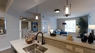 This single-floor residence showcases a thoughtfully designed open-concept layout that connects the kitchen and living areas in a seamless, airy arrangement. The kitchen is a standout feature, equipped with a stainless steel undermount sink, a sleek matte black faucet, and light-toned countertops that extend into a peninsula-style breakfast bar. Decorative pendant lights — including an elegant lantern-style fixture — hang overhead, adding both character and warm illumination to the space. A subway tile backsplash in a neutral tone ties together the kitchen's modern aesthetic. The open living area benefits from a large window that invites abundant natural light, creating a bright and welcoming atmosphere. The living space is comfortably appointed and flows naturally from the kitchen, making it ideal for both everyday living and entertaining. A glimpse into an adjacent bedroom reveals a continuation of the clean, modern design sensibility found throughout the home. Neutral gray walls and contemporary lighting fixtures maintain a cohesive look across all visible spaces. The overall ambiance is modern and functional, with carefully chosen design details that elevate the everyday living experience.