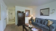 This two-floor home offers a comfortable and traditionally styled living space. The living room serves as a central gathering area, featuring wall-to-wall carpeting in a warm neutral tone that adds softness underfoot. White walls keep the space feeling bright and open, while a framed art piece adds a focal point of color and visual interest. A dark-stained wooden armoire provides substantial storage and anchors one side of the room, complementing a glass-top coffee table and a plush blue sofa adorned with accent pillows. A classic table lamp casts warm ambient light, enhancing the inviting atmosphere. A decorative mirror on the adjacent wall reflects light and adds depth to the room. The open layout allows for easy flow between spaces, with a glimpse into additional rooms visible through the hallway, suggesting a well-connected floor plan. The overall design aesthetic throughout is traditional and homey, with dark wood furnishings contrasting pleasantly against the light walls and carpet, creating a balanced and livable environment across both floors.