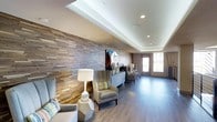 This single-floor space makes a strong first impression with its thoughtfully curated design elements and welcoming ambiance. The most commanding feature is a floor-to-ceiling textured wood-panel accent wall, composed of layered horizontal strips in varying natural tones, adding warmth and visual depth to the interior. Recessed lighting is strategically placed throughout the tray ceiling, casting a soft, even glow that complements the clean white ceiling finish.

The space is furnished with contemporary wingback chairs in neutral upholstery, accented by colorful throw pillows that introduce subtle pops of color. A geometric concrete side table and a classic white table lamp contribute to the modern yet approachable aesthetic. A low-profile dresser-style console serves as a display surface, styled with decorative objects and a table lamp.

Smooth, light-toned hardwood flooring runs throughout, unifying the space and enhancing its sense of openness. Natural light filters in through glass-paneled double doors at the far end, offering a view to the exterior and brightening the room. Overall, the space balances modern design with comfortable, inviting details.