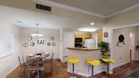 This single-story residence offers a thoughtfully designed layout with interconnected living spaces. The entryway opens into a living area finished with dark hardwood-style flooring and crown molding, with a sliding glass door that draws in natural light and provides access to an outdoor area.

The kitchen is positioned around a peninsula counter topped with granite-style surfaces and features warm wood cabinetry, a full suite of appliances including a refrigerator, microwave, and range, along with a skylight-style ceiling fixture. Vibrant yellow bar stools line the peninsula, adding a pop of color. Adjacent to the kitchen, the dining area accommodates a round wooden table beneath a multi-arm chandelier, complemented by decorative wall accents.

A dedicated laundry room is conveniently accessible directly from the kitchen. The unit includes multiple bedrooms, each equipped with ceiling fans. One bedroom features carpet flooring and an en-suite bathroom, while another offers hardwood-style floors and a walk-in closet. Both bathrooms include warm wood vanities, and one features a shower-tub combination with tile flooring.